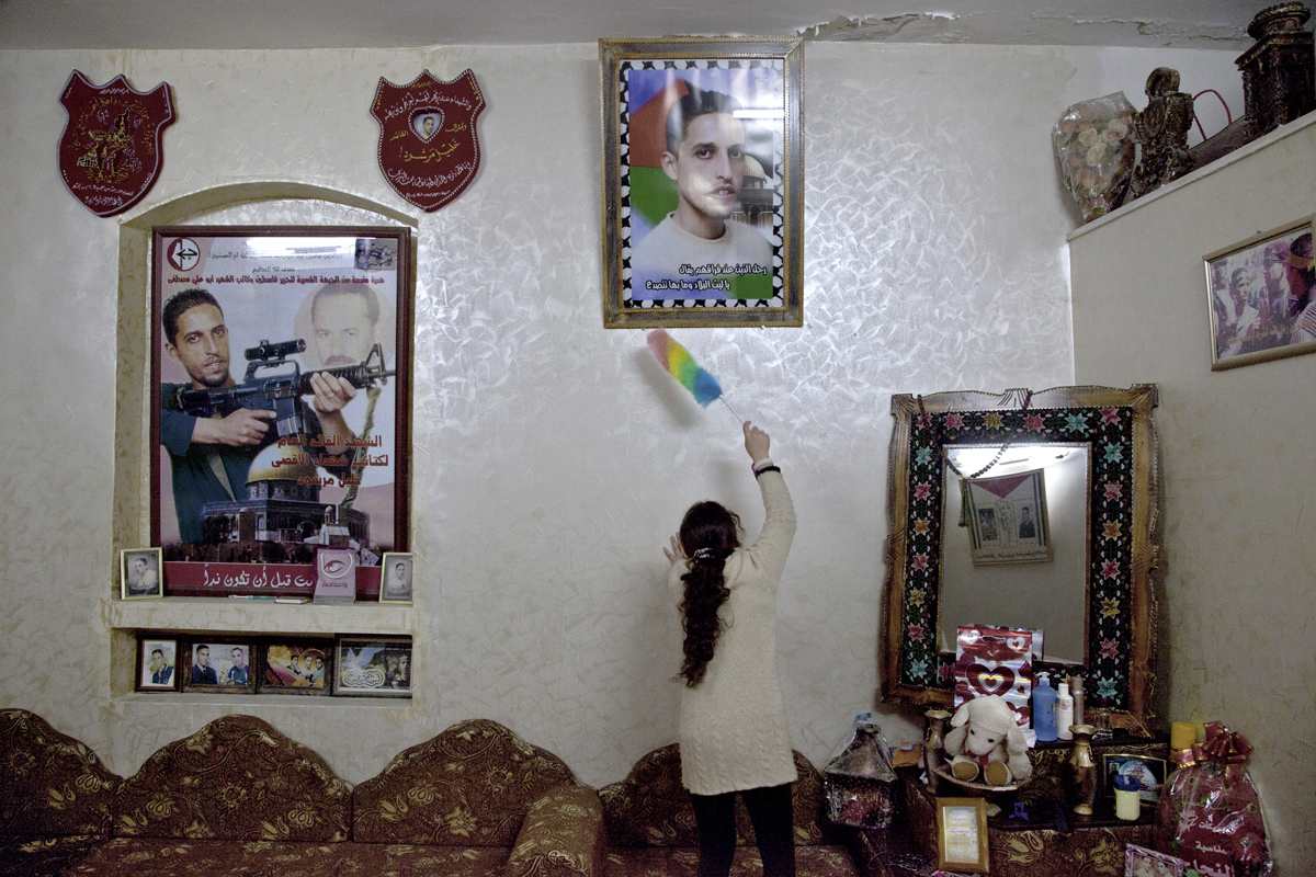 Ahlam Shibli, Sans titre (Death n° 33), Palestine, 2011-2012, tirage chromogénique, 38 x 57 cm Camp de réfugiés de Balata, 16 février 2012 Photos du martyr Khalil Marchoud qu’est en train d’épousseter sa soeur dans le séjour de la maison familiale. Sur l’affiche, cadeau des Brigades Abu Ali Mustafa, il est présenté comme le secrétaire général des Brigades des martyrs d’al-Aqsa à Balata. Courtesy de l’artiste, © Ahlam Shibli